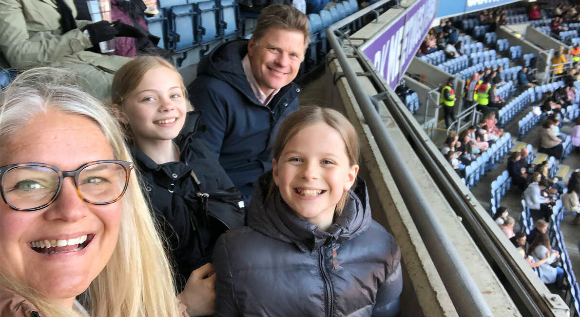 En lykkelig familie på fire, to voksne og to børn, sidder på et stadion. De smiler og kigger på kameraet. Stadionpladserne er delvist fyldte, og nogle personer er synlige i baggrunden.