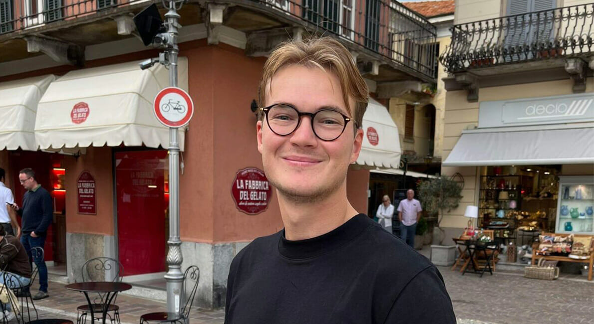 En smilende person med briller står på en brostensbelagt gade foran en café. Caféen har udendørs siddepladser og markiser. I nærheden er der butikker og et cykelforbud. Personen er iført en sort skjorte.