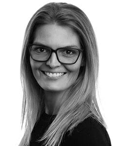 En person med langt hår og briller smiler, mens han ser på kameraet. De har en mørk top på, og billedet er i sort/hvid.