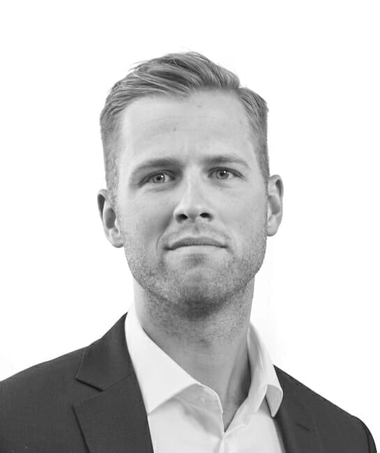 Et gråtonebillede af en person med kort, lyst hår, iført en mørk jakkesæt og en skjorte med hvid krave, og kigger direkte på kameraet mod en almindelig hvid baggrund.