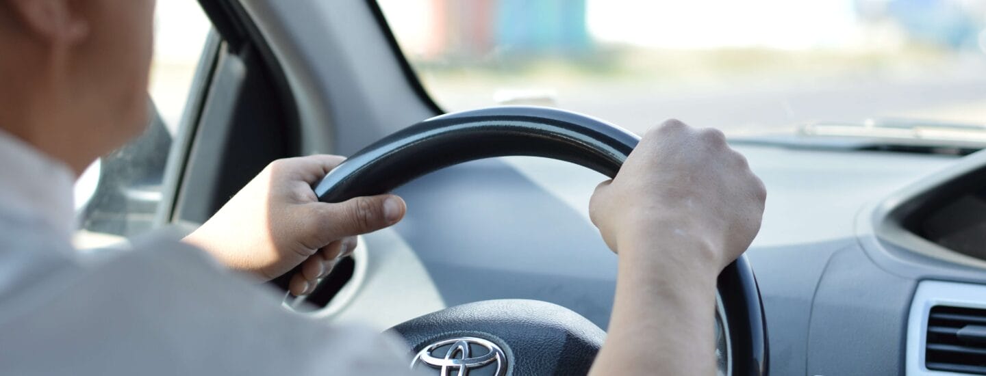 En person i hvid skjorte kører bil og griber solidt om Toyota-rattet. Udsigten fra bagsiden afslører instrumentbrættet og en diset gade bagved, og fanger en scene, der er kendt for mange, der vælger billeje til udlandsdanskere.
