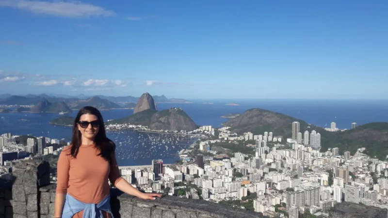 A woman with long hair, wearing sunglasses and an orange shirt, stands smiling in front of a scenic city by the sea. A network of boats dots the bay beneath the famous mountain, all set against a clear blue sky.