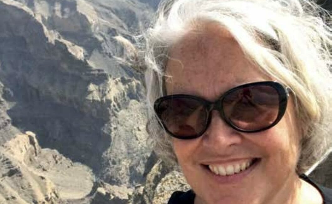 A woman with gray hair and sunglasses smiles while taking a selfie with a background of rugged, rocky cliffs and a deep canyon under a clear sky.