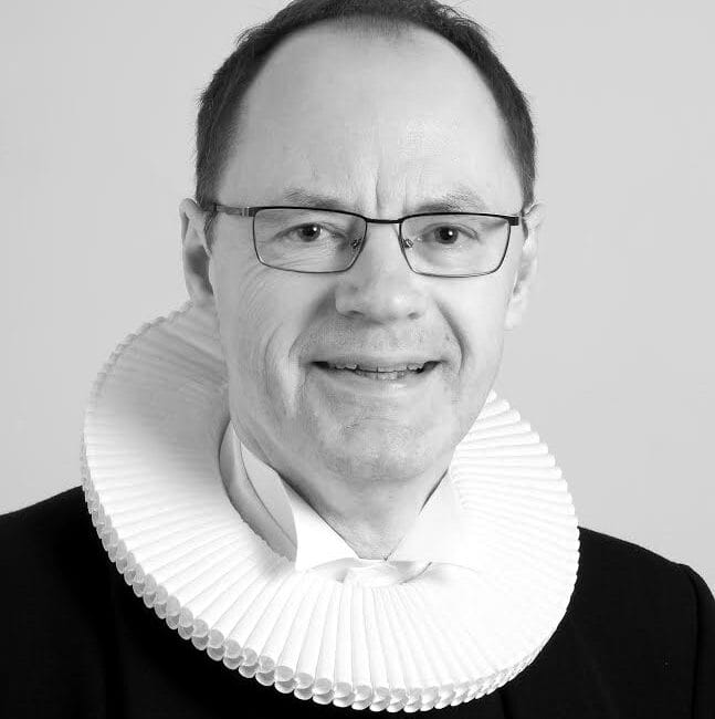 En person, der bærer briller og et mørkt jakkesæt med en traditionel hvid ruff-krave, smiler for et portræt mod en almindelig baggrund.