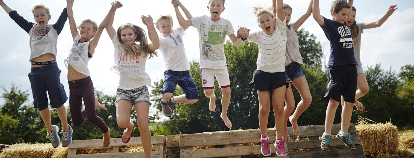A group of nine children are joyfully jumping off a stack of wooden pallets and hay bales outside. Theyre holding hands, with trees and a clear sky in the background, capturing a moment of play and excitement reminiscent of scenes on the Danes Worldwide website.