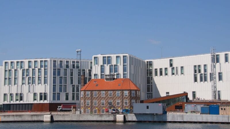 En moderne bygning ved vandet med flere rektangulære vinduer står bag en mindre historisk murstensbygning med rødt tegltag. Scenen er sat mod en klar blå himmel med vand i forgrunden.