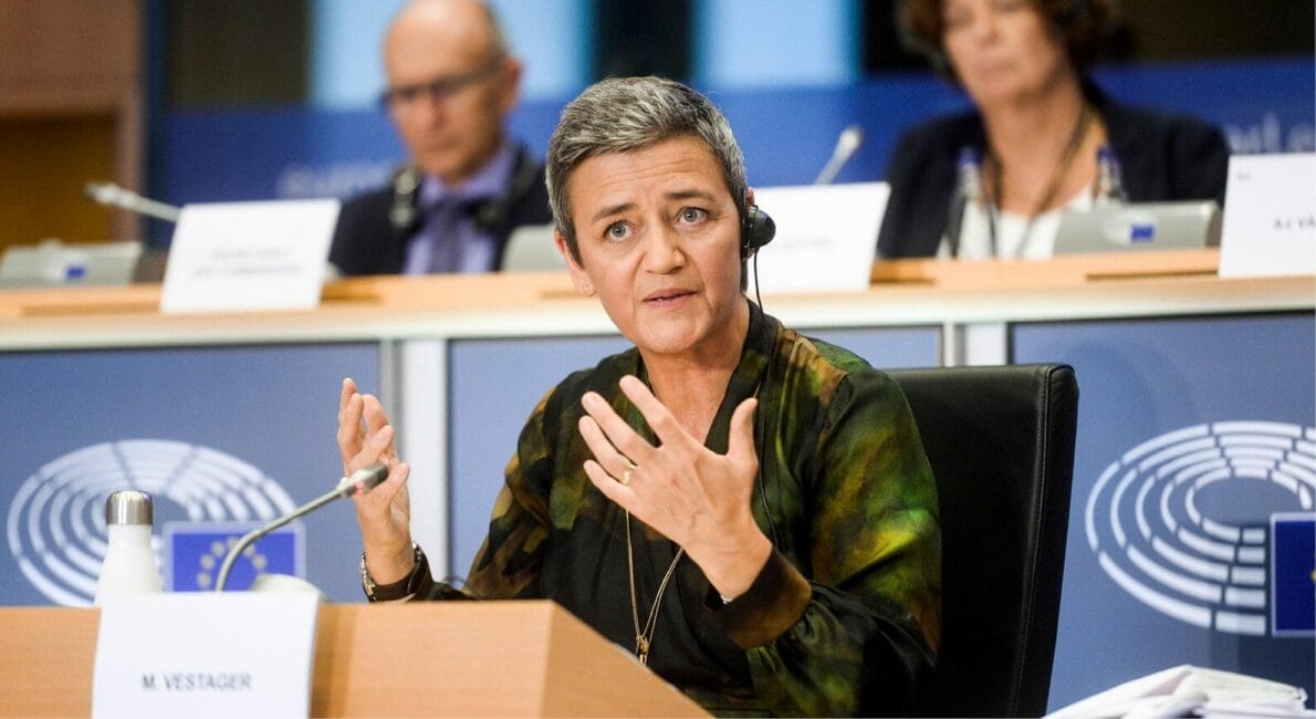En person med kort hår, iført headset og mørkt tøj, taler livligt ved en konference. Siddende foran en mikrofon med "Mister Vestager" på skiltet foran sig, udfylder slørede personer baggrunden.
