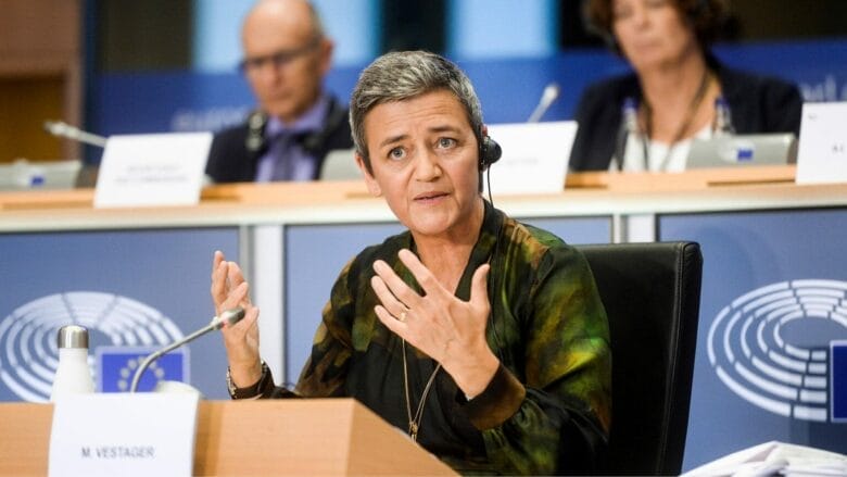 En person med kort hår, iført headset og mørkt tøj, taler livligt ved en konference. Siddende foran en mikrofon med "Mister Vestager" på skiltet foran sig, udfylder slørede personer baggrunden.