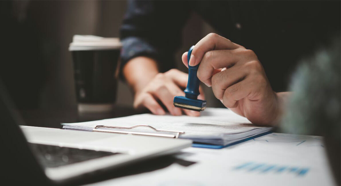 En person holder et gummistempel over et dokument på et skrivebord. En udklipsholder med papirer, en bærbar computer og en kaffekop er i nærheden. Indstillingen foreslår et kontormiljø med fokus på administrative eller godkendelsesaktiviteter.