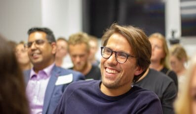 En gruppe mennesker sidder i et lokale, muligvis samlet til et Danes Worldwide seminar. En mand i forgrunden med briller og skæg smiler, mens andre omkring ham sidder opmærksomt og absorberer præsentationen.